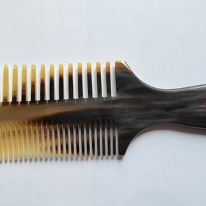 Könnte beinhalten: Ein schwarzer und brauner Hornkamm mit einem Design mit breiten Zähnen. Der Kamm hat einen langen Griff und ein abgerundetes Ende.
