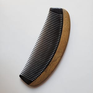 May include: A hair comb featuring a curved design with a smooth, dark brown wooden handle on one side and a series of fine, black teeth on the other. The comb has a polished finish, showcasing the natural grain of the wood and the solid black color.