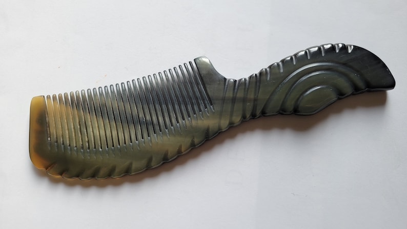 May include: A handcrafted comb with a dark gray and tan color scheme. The comb features closely spaced teeth and a decorative handle with a swirling design. The comb is made of a natural material.