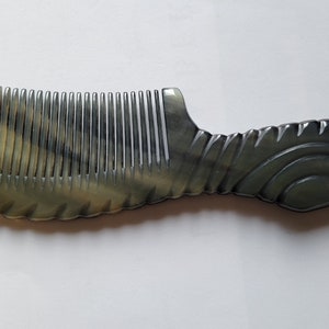 May include: A handcrafted comb with a unique design. The comb features a handle with a curved, textured pattern and a row of evenly spaced teeth. The color is a mix of light and dark tones, creating a marbled effect. The comb is made of a durable material.