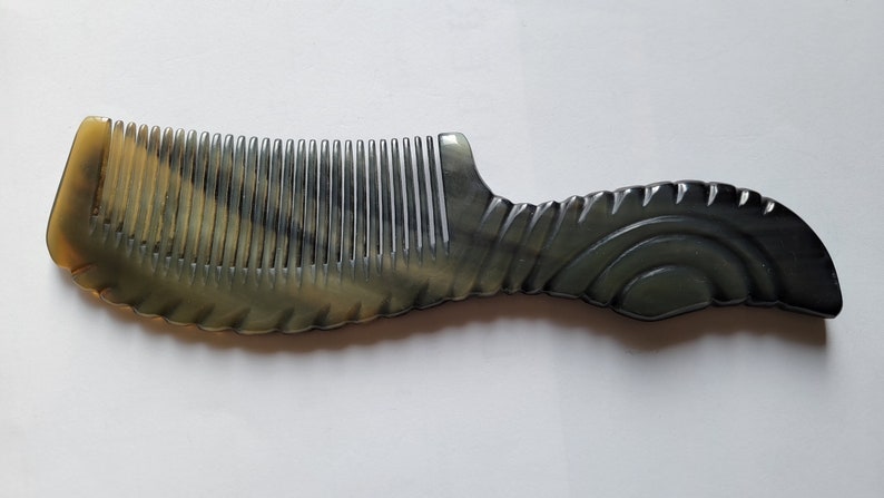 May include: A handcrafted comb with a unique design. The comb features a handle with a curved, textured pattern and a row of evenly spaced teeth. The color is a mix of light and dark tones, creating a marbled effect. The comb is made of a durable material.