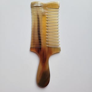 K&ouml;nnte beinhalten: Ein doppelseitiger Haarkamm aus nat&uuml;rlichem Horn mit einem glatten Griff und zwei verschiedenen Zahnreihen unterschiedlicher Breite. Das Material weist ein durchscheinendes, marmoriertes Muster in Bernstein-, Braun- und Cremet&ouml;nen auf.