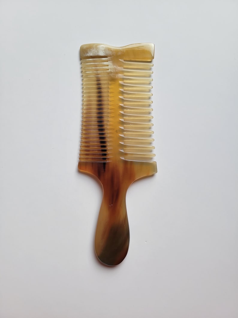 K&ouml;nnte beinhalten: Ein doppelseitiger Haarkamm aus nat&uuml;rlichem Horn mit einem glatten Griff und zwei verschiedenen Zahnreihen unterschiedlicher Breite. Das Material weist ein durchscheinendes, marmoriertes Muster in Bernstein-, Braun- und Cremet&ouml;nen auf.