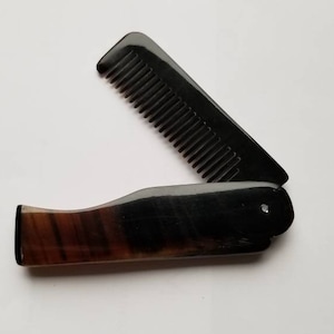 May include: A black folding comb with a handle featuring a brown and black pattern. The comb is open, revealing the teeth. The design is sleek and compact, suitable for travel or everyday use.