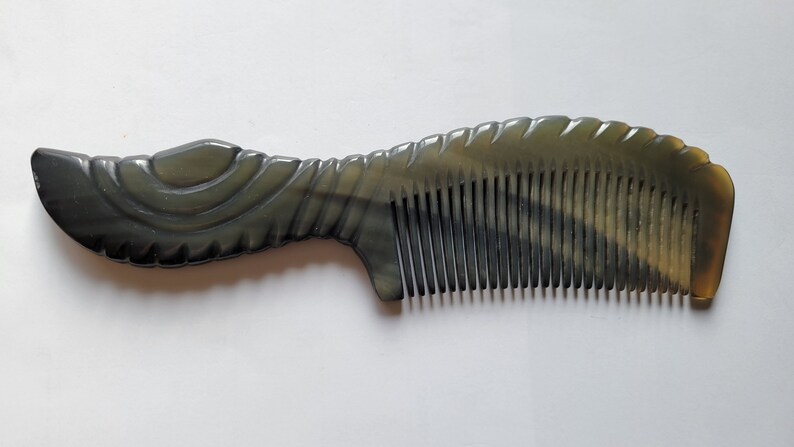 May include: A dark gray comb with a decorative handle and fine teeth. The comb has a curved design and is made of a smooth, polished material. The handle features carved details, and the teeth are evenly spaced.
