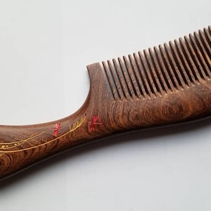 May include: A wooden comb with a brown and red design. The comb has a long handle with a curved end. The handle is decorated with a gold design of leaves and a dragonfly.