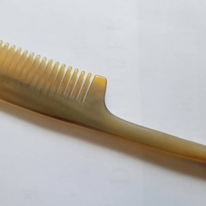 Könnte beinhalten: Ein hellbrauner Kamm mit langem Griff und eng stehenden Zähnen. Der Kamm hat eine gebogene Form und eine glatte, polierte Oberfläche. Der Griff verjüngt sich zu einer Spitze, und die Zähne sind gleichmäßig verteilt.