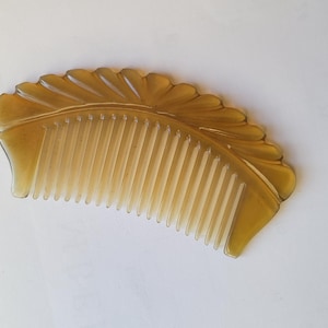 May include: A light amber-colored comb with a decorative, scalloped edge. The comb's teeth are closely spaced and run the length of the curved design. The comb is made of a translucent material, and is likely used for hair care.