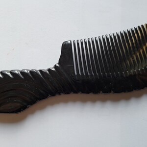 May include: A dark brown comb with a curved handle and closely spaced teeth. The handle has a decorative, textured design. The comb's color transitions to a lighter, translucent brown at the top.