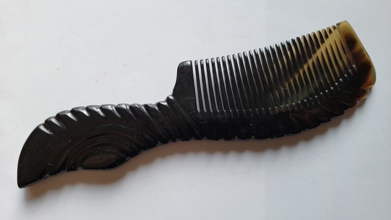 May include: A dark brown comb with a curved handle and closely spaced teeth. The handle has a decorative, textured design. The comb's color transitions to a lighter, translucent brown at the top.