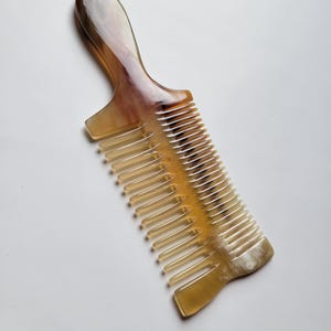 K&ouml;nnte beinhalten: Ein Haarkamm aus poliertem Horn mit einem glatten, geschwungenen Griff und einem zweigeteilten Design. Das Material zeigt einen nat&uuml;rlichen, durchscheinenden Farbverlauf in Bernstein-, Creme- und Dunkelbraunt&ouml;nen mit sichtbaren organischen Maserungen.
