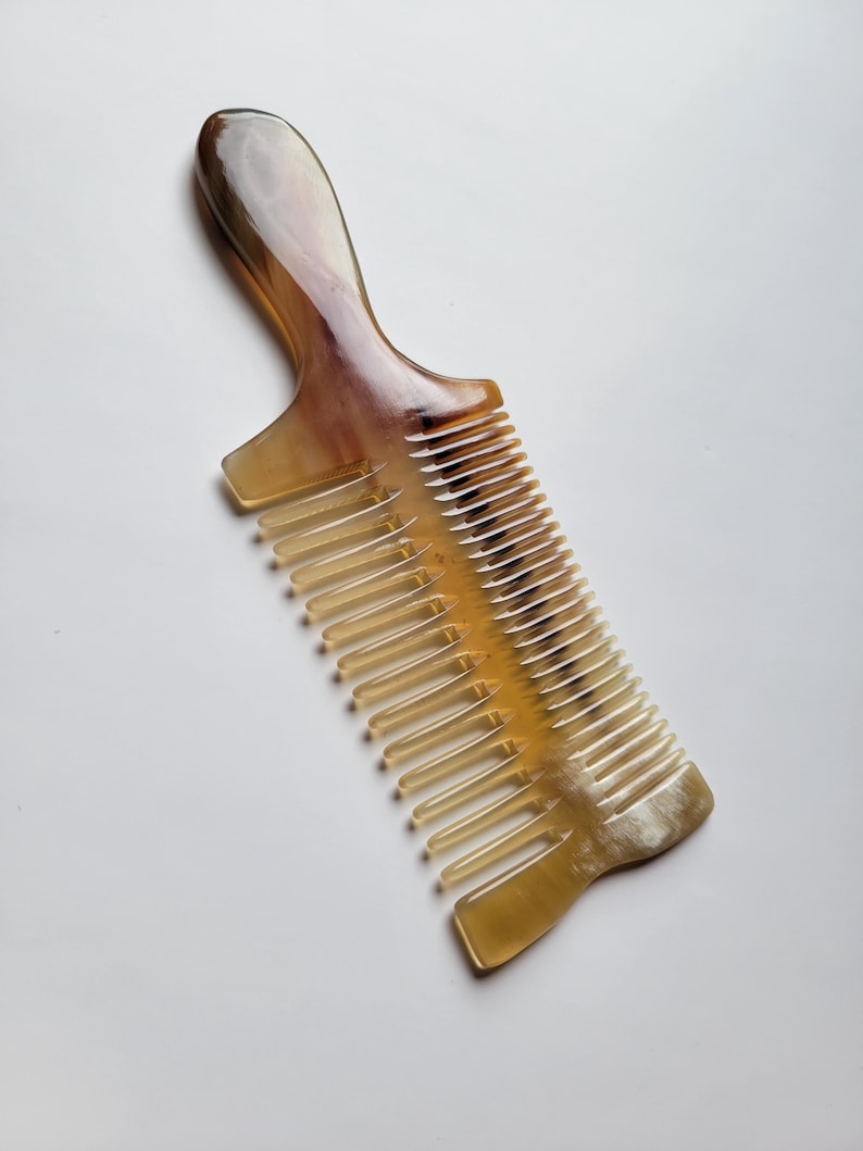 K&ouml;nnte beinhalten: Ein Haarkamm aus poliertem Horn mit einem glatten, geschwungenen Griff und einem zweigeteilten Design. Das Material zeigt einen nat&uuml;rlichen, durchscheinenden Farbverlauf in Bernstein-, Creme- und Dunkelbraunt&ouml;nen mit sichtbaren organischen Maserungen.
