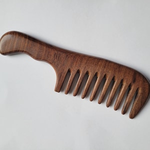 May include: A dark brown wooden hair comb with a smooth, polished finish and a curved handle. The comb features wide, evenly spaced teeth designed for detangling, with a visible natural wood grain pattern throughout the entire structure.