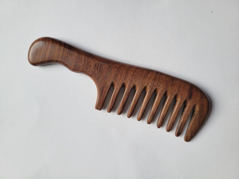 May include: A dark brown wooden hair comb with a smooth, polished finish and a curved handle. The comb features wide, evenly spaced teeth designed for detangling, with a visible natural wood grain pattern throughout the entire structure.
