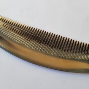 May include: A natural horn comb with a curved shape and fine teeth. The comb has a gradient of colors, from light tan to dark brown. The comb is approximately 6 inches long.
