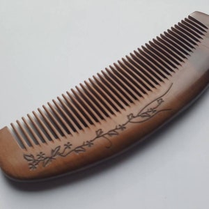 May include: A wooden comb with fine teeth and a curved handle. The comb is a warm brown color and features an etched floral design along the handle. The comb is designed for hair care and grooming.