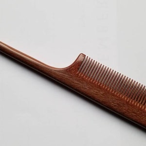 A long, thin, wooden comb with a pointed end and a narrow, rectangular comb section. The comb is made of a reddish-brown wood with a natural grain pattern.