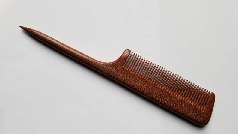 A long, thin, wooden comb with a pointed end and a narrow, rectangular comb section. The comb is made of a reddish-brown wood with a natural grain pattern.