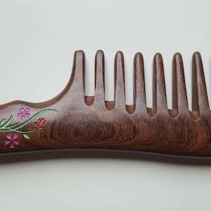 A brown wooden comb with a floral design slightly carved into the handle. The flowers are pink, green, and white. This wood comb has extra wide teeth and is good for curly, long, and thick hair.