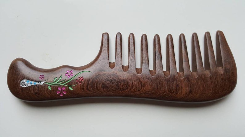 A brown wooden comb with a floral design slightly carved into the handle. The flowers are pink, green, and white. This wood comb has extra wide teeth and is good for curly, long, and thick hair.