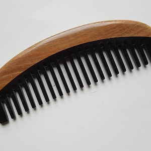 Puede incluir: Un peine de madera con una sección de peine de plástico negro. El peine es curvo y tiene un acabado de madera natural.