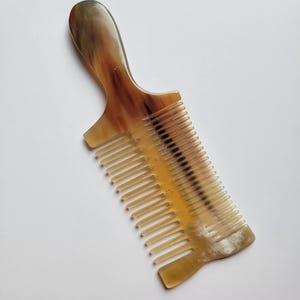 K&ouml;nnte beinhalten: Ein Haarkamm mit Griff aus poliertem Hornmaterial. Der Kamm hat ein durchscheinendes, marmoriertes Aussehen mit vermischten Farbt&ouml;nen in Bernstein, Creme und Dunkelbraun. Er verf&uuml;gt &uuml;ber eine Reihe feiner, gleichm&auml;&szlig;ig angeordneter Zinken.