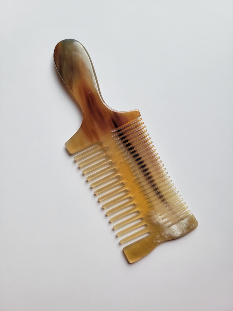 K&ouml;nnte beinhalten: Ein Haarkamm mit Griff aus poliertem Hornmaterial. Der Kamm hat ein durchscheinendes, marmoriertes Aussehen mit vermischten Farbt&ouml;nen in Bernstein, Creme und Dunkelbraun. Er verf&uuml;gt &uuml;ber eine Reihe feiner, gleichm&auml;&szlig;ig angeordneter Zinken.