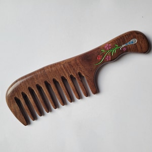 May include: A dark brown wooden hair comb with a curved handle. The handle features an inlaid design of a green stem with small pink and red flowers and a silver-toned accent piece with blue detailing near the top of the handle.