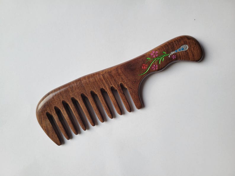 May include: A dark brown wooden hair comb with a curved handle. The handle features an inlaid design of a green stem with small pink and red flowers and a silver-toned accent piece with blue detailing near the top of the handle.