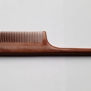 A long, wooden comb with a brown, reddish hue. The comb has a long, thin handle and  a slim, rectangular comb section. it's designed for hair styling and sectioning.