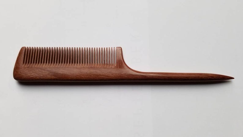 A long, wooden comb with a brown, reddish hue. The comb has a long, thin handle and  a slim, rectangular comb section. it's designed for hair styling and sectioning.