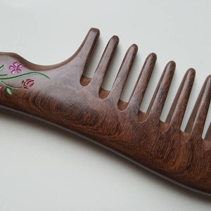 May include: A dark brown wooden hair comb featuring a curved handle and wide teeth. The handle is decorated with an inlaid floral design consisting of small pink flowers, green stems, and a silver-toned accent piece with blue and white details.