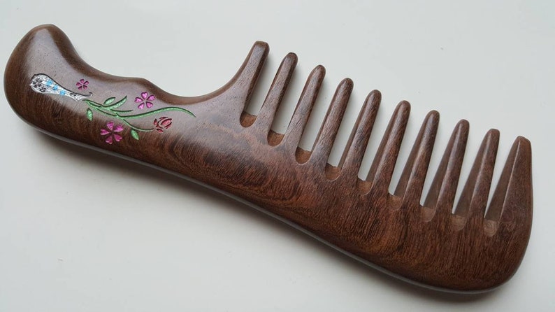May include: A dark brown wooden hair comb featuring a curved handle and wide teeth. The handle is decorated with an inlaid floral design consisting of small pink flowers, green stems, and a silver-toned accent piece with blue and white details.