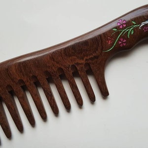May include: A dark brown wooden hair comb with a wide-tooth design and a curved handle. The handle features an inlaid floral motif with pink flower petals, green stems, and a blue and white bird-like figure crafted from iridescent shell material.