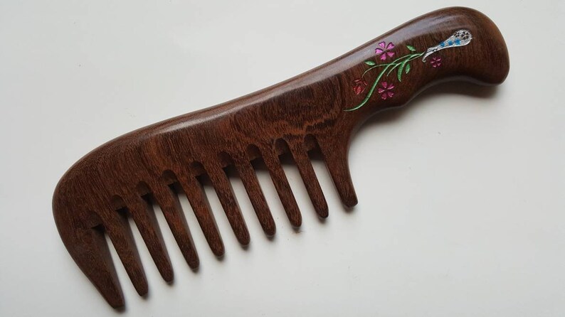 May include: A dark brown wooden hair comb with a wide-tooth design and a curved handle. The handle features an inlaid floral motif with pink flower petals, green stems, and a blue and white bird-like figure crafted from iridescent shell material.