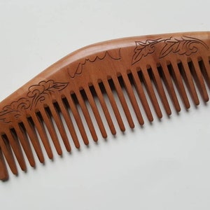 May include: A wooden comb with a curved handle and fine teeth. The comb is a warm brown color and features an engraved floral design along the top edge. The comb is designed for hair care and styling.