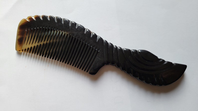 May include: A dark brown comb with a curved handle and fine teeth. The comb has a decorative, carved handle and a lighter brown tip. The comb is made of a smooth, polished material.