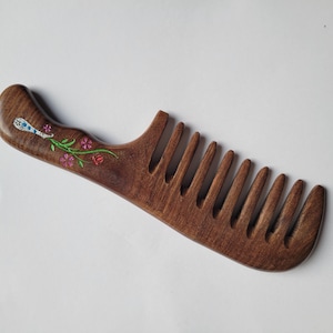 May include: A dark brown wooden hair comb with a curved handle featuring an inlaid floral design. The inlay consists of a light blue and white patterned element, green stems, and small pink flowers embedded into the wood surface.