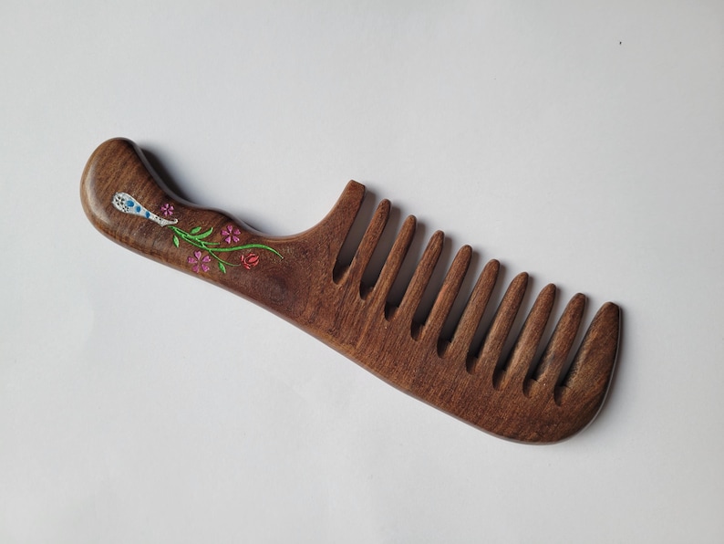 May include: A dark brown wooden hair comb with a curved handle featuring an inlaid floral design. The inlay consists of a light blue and white patterned element, green stems, and small pink flowers embedded into the wood surface.