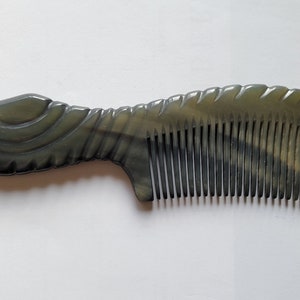 May include: A dark gray comb with a curved handle and fine teeth. The handle has a decorative, carved design. The comb is made of a smooth, polished material and is set against a white background.