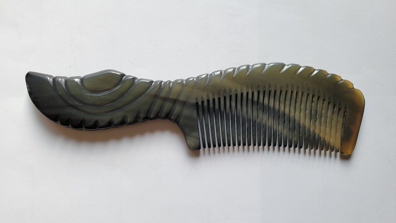 May include: A dark gray comb with a curved handle and fine teeth. The handle has a decorative, carved design. The comb is made of a smooth, polished material and is set against a white background.
