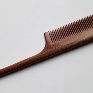 A long, thin, wooden comb with a pointed handle. The comb is made of dark brown pau rosa wood and has fine teeth.  it is designed for precision sectioning and hair styling.
