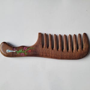 May include: A dark brown wooden comb with a wide-tooth design and a curved handle. The handle features a painted floral motif with small pink and red flowers, green stems, and a white decorative element with blue dots on the left side.
