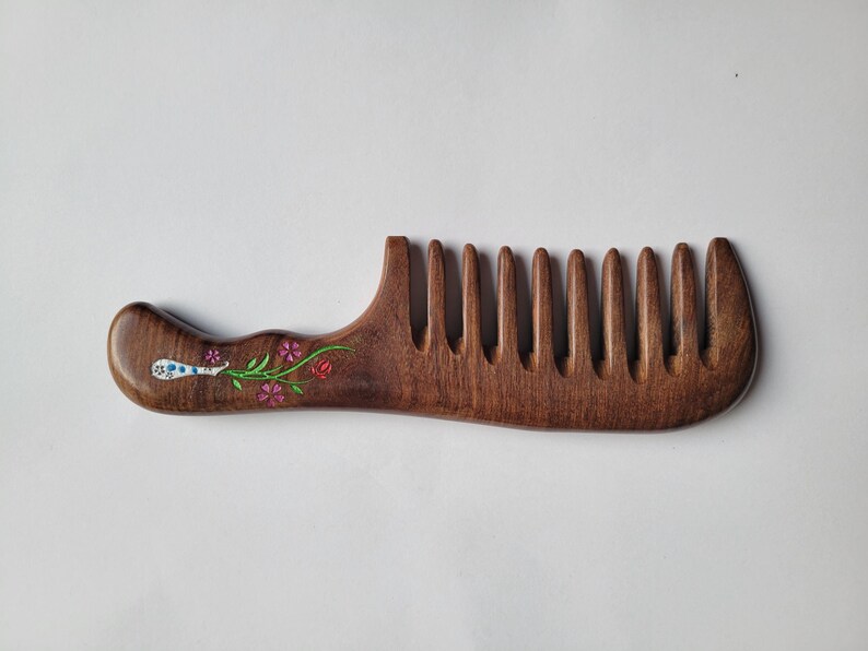 May include: A dark brown wooden comb with a wide-tooth design and a curved handle. The handle features a painted floral motif with small pink and red flowers, green stems, and a white decorative element with blue dots on the left side.