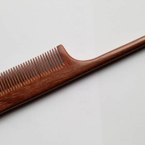 A brown wooden comb with a long, thin handle. The comb has a rounded edge and a pointed tip.