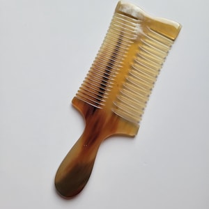 K&ouml;nnte beinhalten: Ein Haarkamm mit Griff, der ein durchscheinendes, marmoriertes Muster in Bernstein-, Braun- und Cremet&ouml;nen aufweist. Der Kamm hat einen rechteckigen Kopf mit feinen, gleichm&auml;&szlig;ig angeordneten Zinken und einen glatten, abgerundeten Griff.