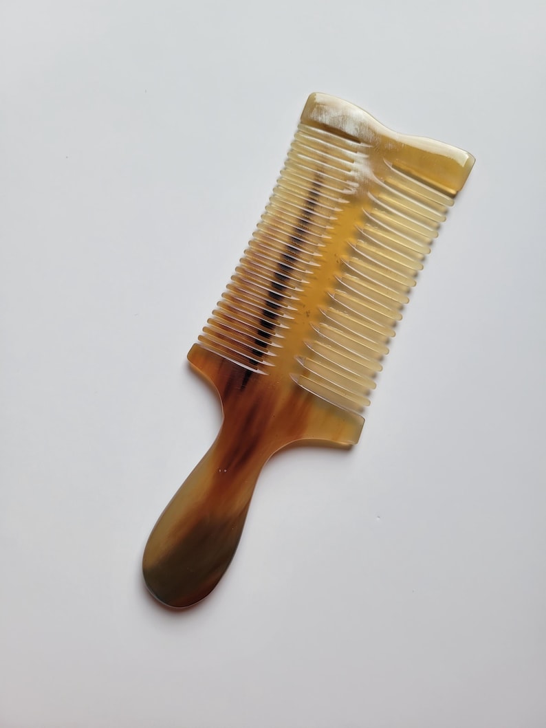K&ouml;nnte beinhalten: Ein Haarkamm mit Griff, der ein durchscheinendes, marmoriertes Muster in Bernstein-, Braun- und Cremet&ouml;nen aufweist. Der Kamm hat einen rechteckigen Kopf mit feinen, gleichm&auml;&szlig;ig angeordneten Zinken und einen glatten, abgerundeten Griff.