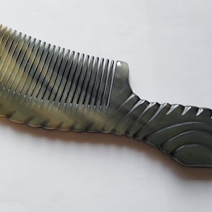 May include: A horn comb with a dark gray and tan color scheme. The comb has fine teeth and a decorative handle with a curved design. The handle has a series of ridges and a curved shape.