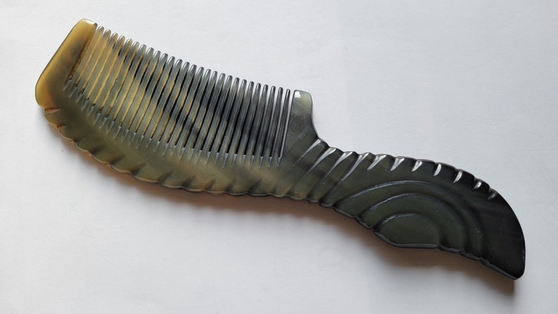 May include: A horn comb with a dark gray and tan color scheme. The comb has fine teeth and a decorative handle with a curved design. The handle has a series of ridges and a curved shape.