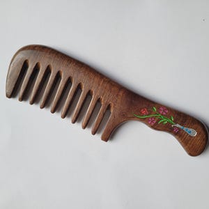 May include: A dark brown wooden wide-tooth comb featuring a smooth, curved handle. The handle is decorated with an inlaid design of a silver-toned vase containing a small green stem with pink and red flowers and blue accents.